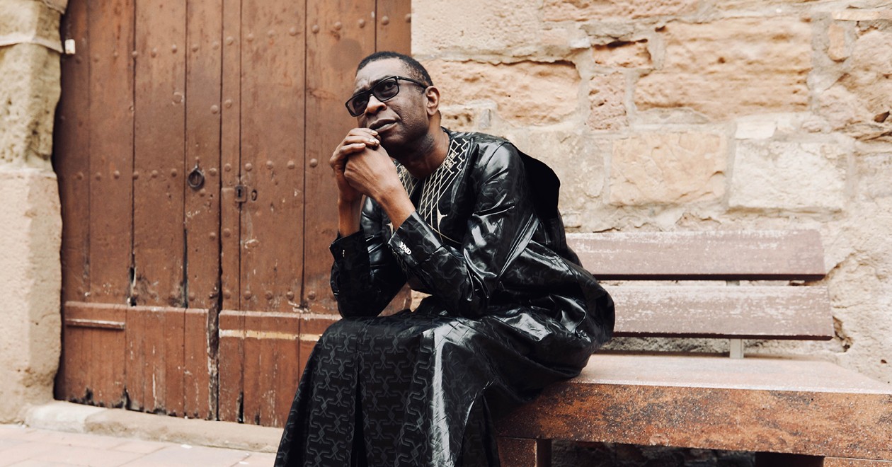 Youssou N'Dour assis sur un banc devant une porte en bois ancienne, vêtu d'un boubou noir.