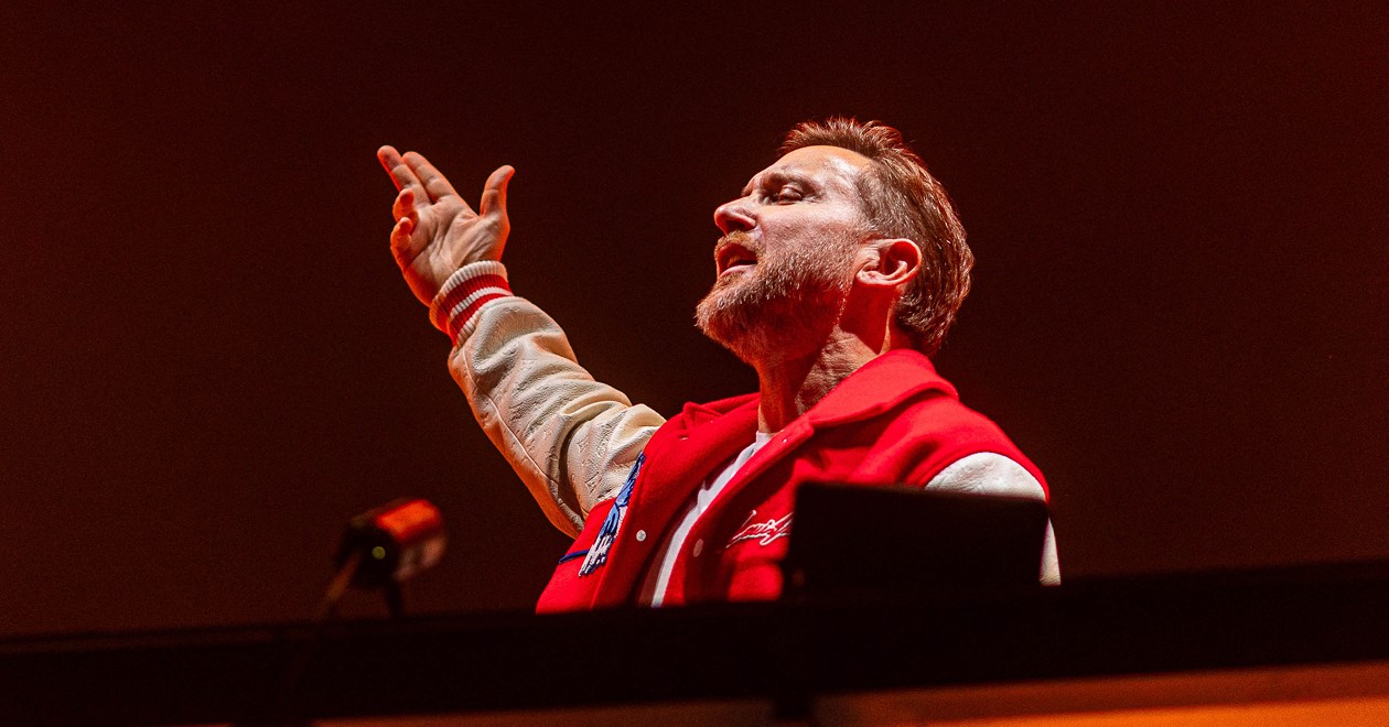 David Guetta at the decks during a live performance, wearing a red jacket, arm raised