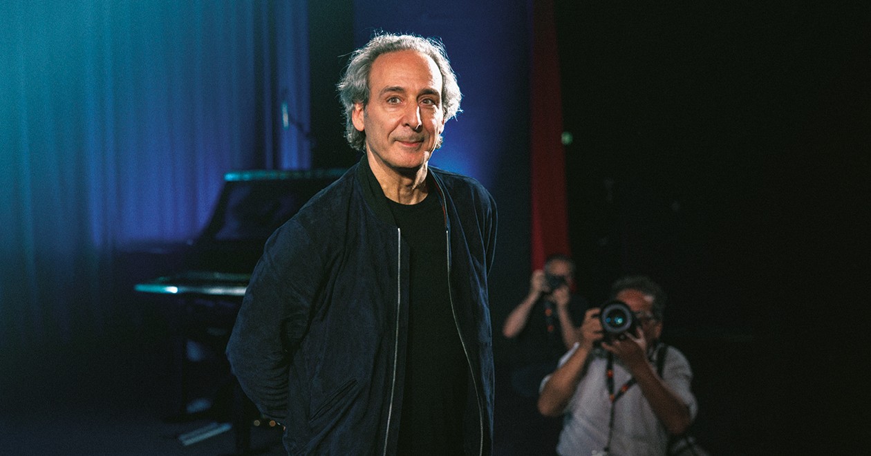 Alexandre Desplat on stage, smiling, in front of a grand piano with a photographer in the background