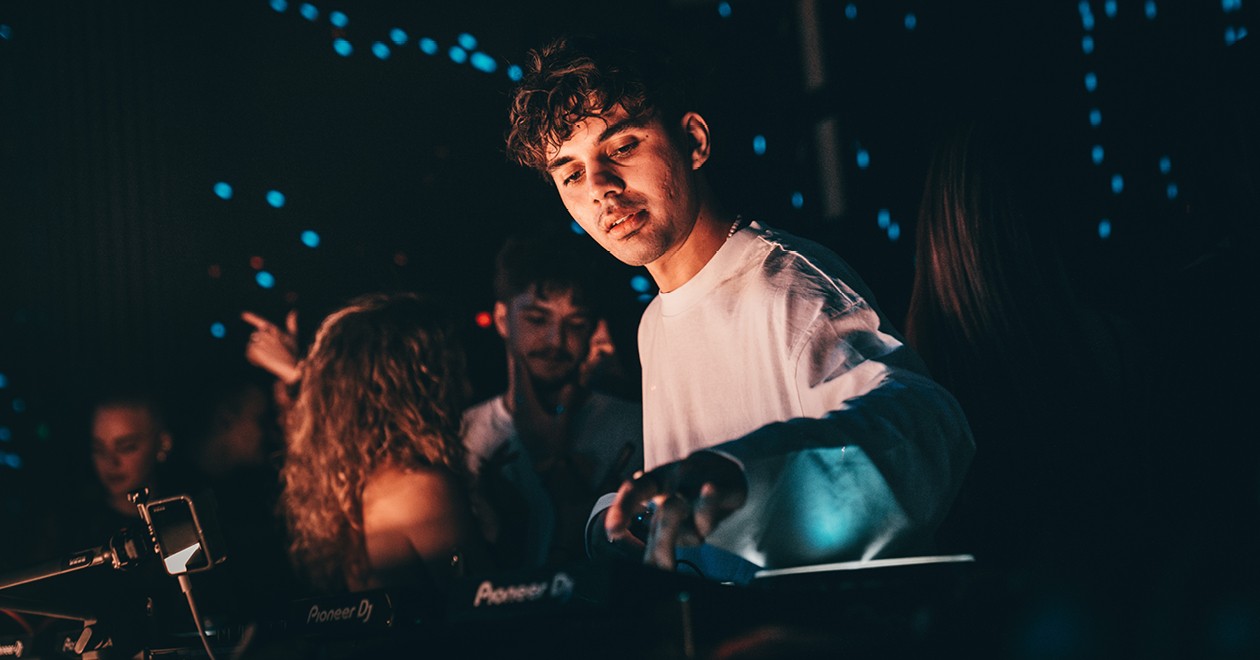 Rivo at the decks during an event, under blue lights in a concert venue